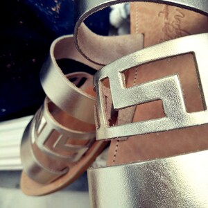 May include: Gold leather sandals with a cut-out design. The sandals have a thick strap and a flat sole.