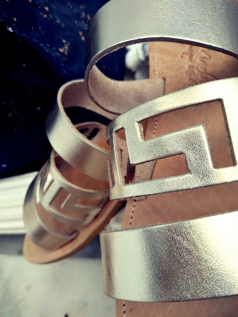 May include: Gold leather sandals with a cut-out design. The sandals have a thick strap and a flat sole.