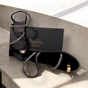 May include: A pair of black sandals with a sparkly design. The sandals have a thin strap that wraps around the ankle and a toe ring. The sandals are on a white surface with a black box that says "Marbe Sandals".