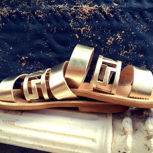 May include: Gold leather sandals with a geometric cut-out design. The sandals have a brown leather sole and are resting on a white surface.