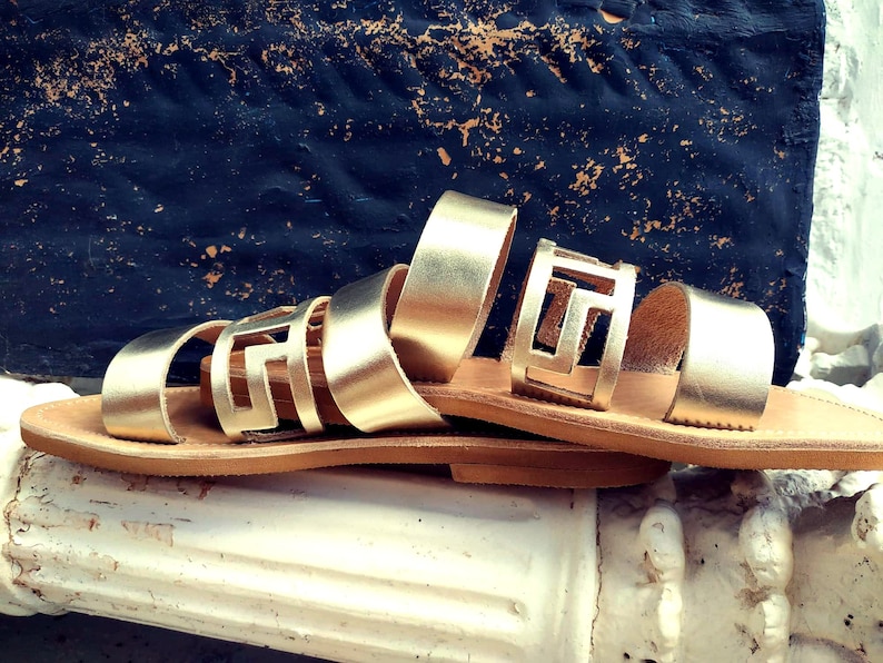 May include: Gold leather sandals with a geometric cut-out design. The sandals have a brown leather sole and are resting on a white surface.