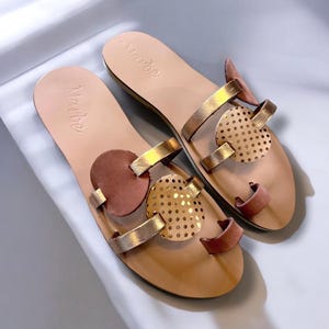 May include: A pair of gold leather sandals with brown leather accents. The sandals have a geometric design with circles and a perforated circle. The sandals are labelled "Marloe".