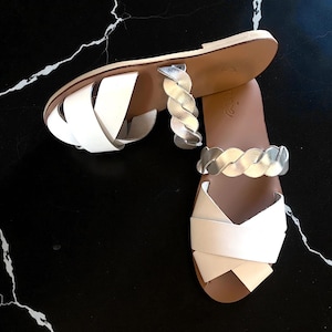 May include: A pair of white leather sandals with silver braided straps. The sandals have a flat sole and a crisscross design on the top. The sandals are displayed on a black marble surface.