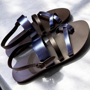 May include: A pair of brown leather sandals with metallic blue straps. The sandals have a flat sole and a buckle closure on the ankle strap. The sandals are designed for casual wear.