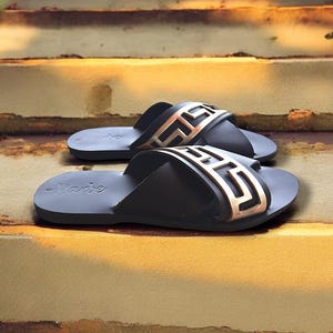 May include: A pair of black leather sandals with a gold-coloured Greek key design across the strap. The sandals are on a set of concrete steps.