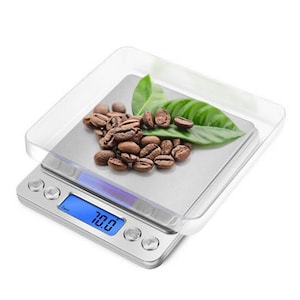 May include: A silver digital kitchen scale with a clear plastic tray, displaying 1984 grams. The scale has three buttons and a blue digital display. The tray holds roasted coffee beans and green leaves.