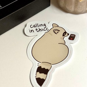 May include: A sticker featuring a cartoon raccoon holding a phone with the text "WORK" on the screen. The raccoon is sitting with its legs crossed and has a speech bubble above its head that says "calling in thicc".