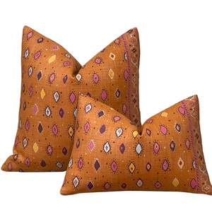 May include: Two decorative throw pillows with a warm orange background and a repeating pattern of diamonds in pink, yellow, and dark purple. The pillows have a textured appearance and a decorative border.