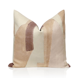 May include: Square cushion with an abstract design in beige, tan, and brown hues. The cushion features a geometric pattern with curved and rectangular shapes, creating a modern look. The cushion is made of a textured fabric.