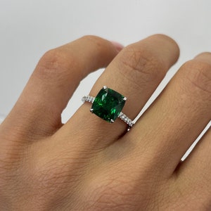 Op de afbeelding: Een zilveren ring met een grote, smaragdgroene, kussen geslepen edelsteen, omringd door kleinere, heldere edelstenen.