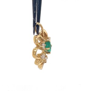 Puede incluir: Un colgante de oro con una piedra central de esmeralda ovalada y peque&ntilde;os detalles de diamantes. El colgante tiene un dise&ntilde;o decorativo y calado. El colgante est&aacute; sujeto por una herramienta negra sobre un fondo blanco.
