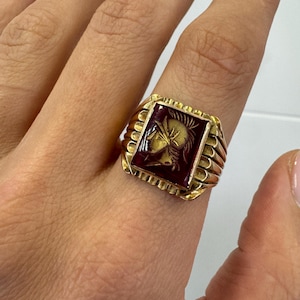 Puede incluir: Un anillo de oro con una piedra roja que presenta una imagen tallada de la cabeza de un guerrero.
