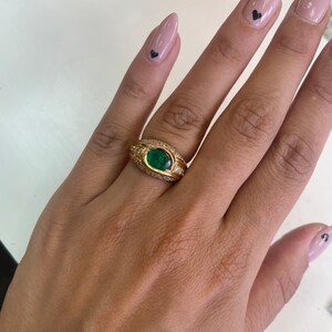 K&ouml;nnte beinhalten: Ein goldener Ring mit einem gro&szlig;en smaragdgr&uuml;nen Edelstein, der von kleineren Diamanten umgeben ist. Der Ring wird an einem Finger getragen.