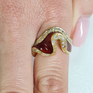 May include: A gold ring with a red enamel design and clear cubic zirconia stones. The ring is shaped like a wave and has a unique, modern design.