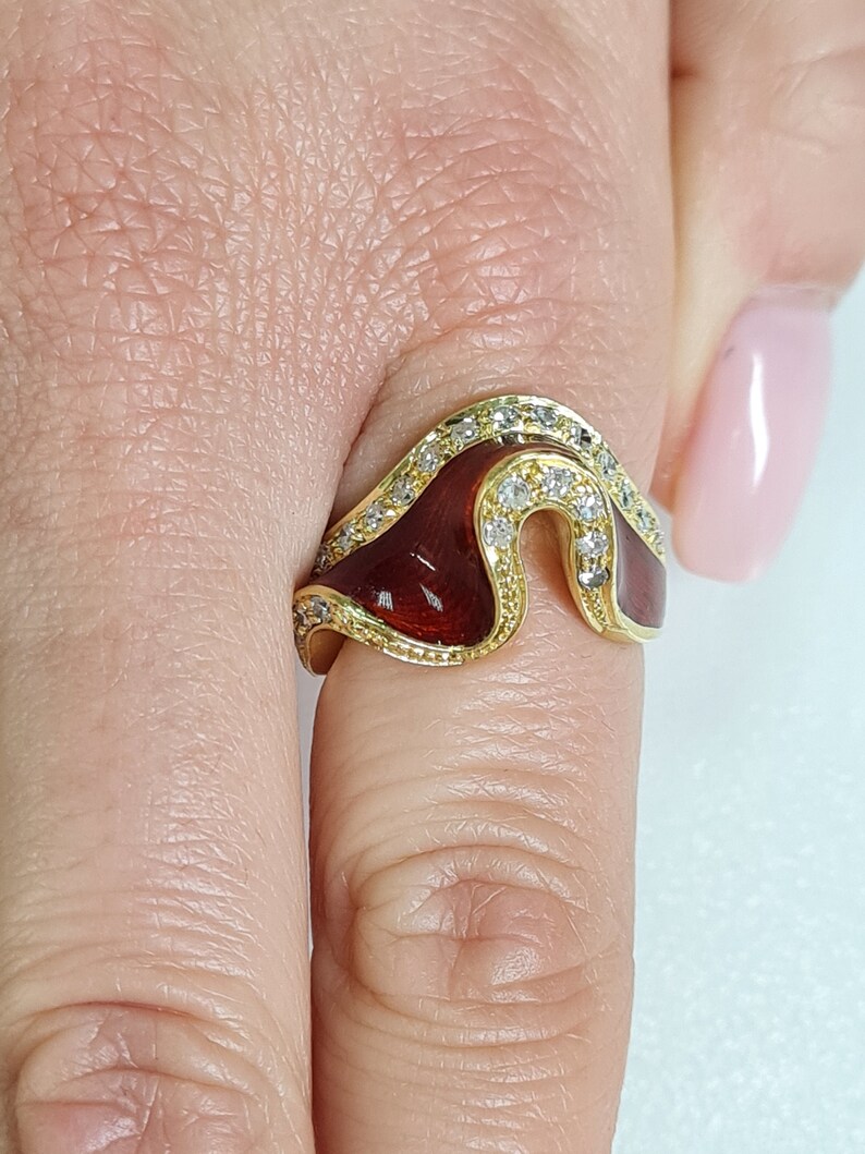 May include: A gold ring with a red enamel design and clear cubic zirconia stones. The ring is shaped like a wave and has a unique, modern design.