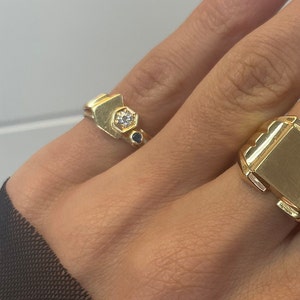 May include: Two gold rings on a hand. The ring on the index finger has a geometric design with a small white stone and two blue stones. The ring on the middle finger is a plain gold signet ring.
