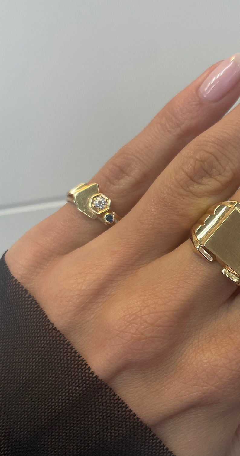 May include: Two gold rings on a hand. The ring on the index finger has a geometric design with a small white stone and two blue stones. The ring on the middle finger is a plain gold signet ring.