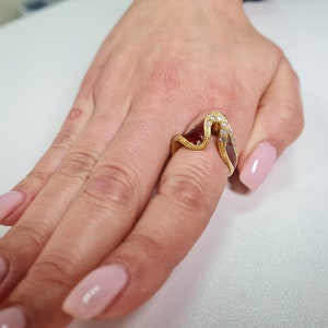 May include: A gold ring with a wave-like design and clear stones. The ring is on a finger with pink nail polish.
