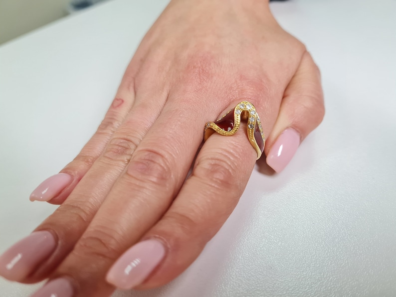 May include: A gold ring with a wave-like design and clear stones. The ring is on a finger with pink nail polish.