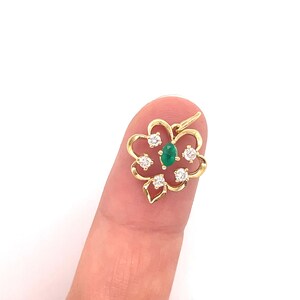 Puede incluir: Un colgante dorado con forma de flor estilizada. El colgante presenta una piedra preciosa verde ovalada central, rodeada de cinco piedras preciosas redondas y claras. El colgante est&aacute; sujeto por un dedo sobre un fondo blanco.