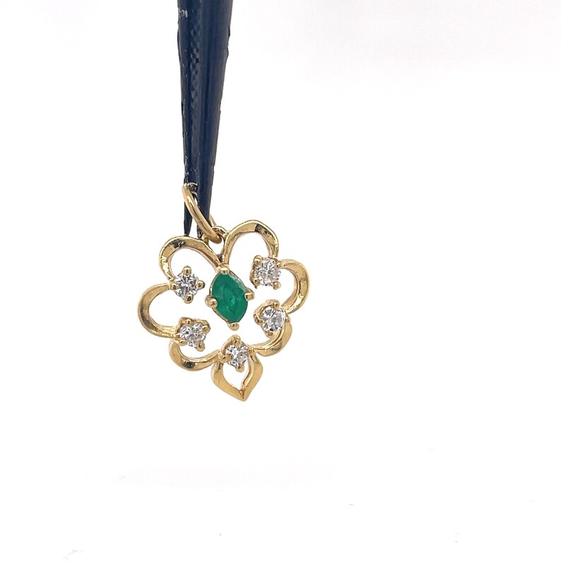 Puede incluir: Colgante de oro con dise&ntilde;o floral, con una gema verde central tallada en esmeralda y cuatro gemas claras circundantes. El colgante est&aacute; suspendido de un cord&oacute;n negro sobre un fondo blanco. El intrincado dise&ntilde;o del colgante a&ntilde;ade elegancia.