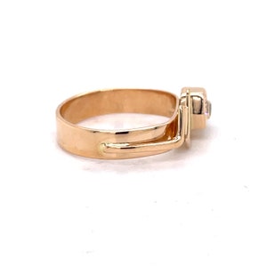 May include: A gold ring with a single clear stone set in a square bezel. The ring has a unique design with a band that wraps around the finger and then extends to the side, creating a unique and modern look.