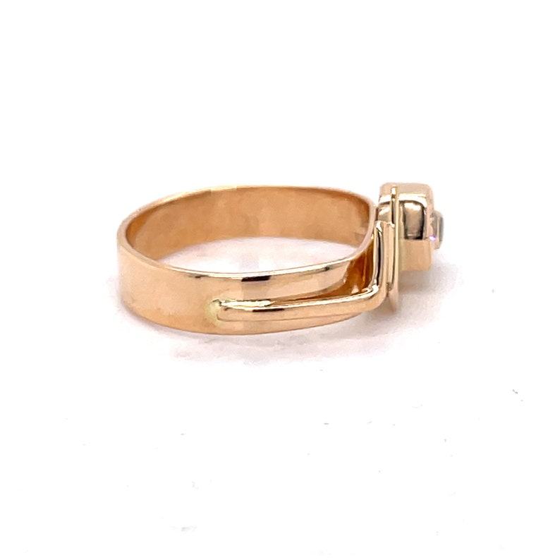 May include: A gold ring with a single clear stone set in a square bezel. The ring has a unique design with a band that wraps around the finger and then extends to the side, creating a unique and modern look.