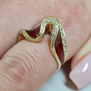May include: A gold ring with a red enamel design and clear stones. The ring is shaped like a wave and has a delicate, intricate design.