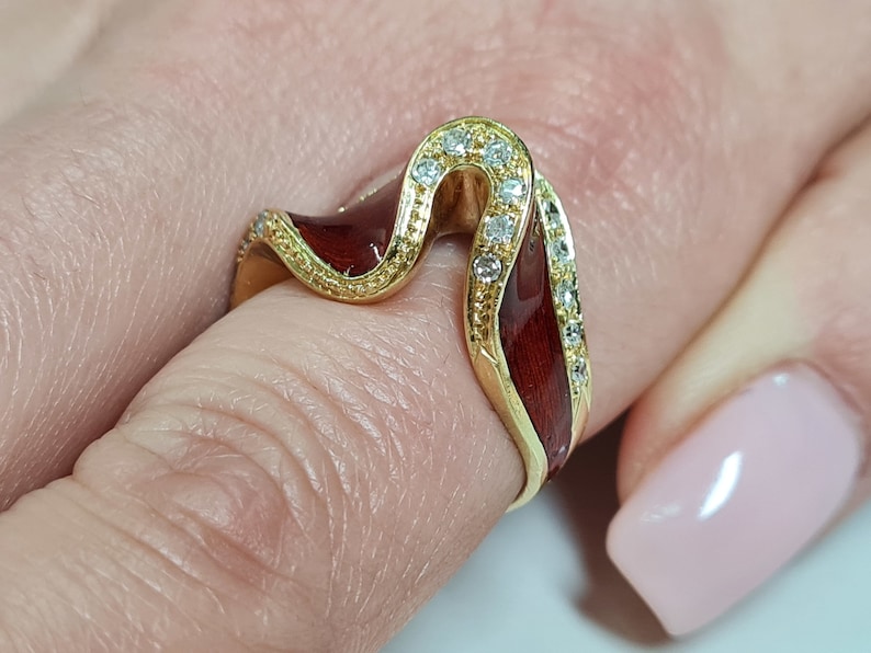 May include: A gold ring with a red enamel design and clear stones. The ring is shaped like a wave and has a delicate, intricate design.