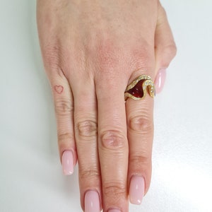 May include: A gold ring with a red gemstone and a wave-like design. The ring is set with small diamonds.