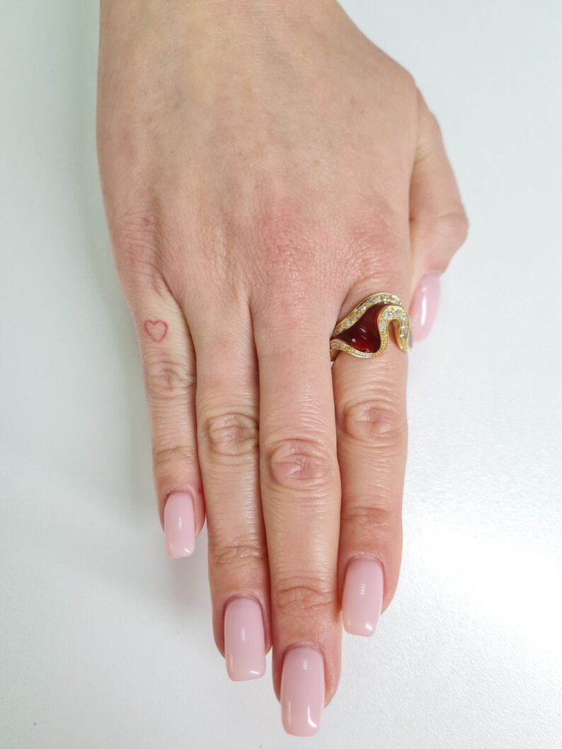 May include: A gold ring with a red gemstone and a wave-like design. The ring is set with small diamonds.
