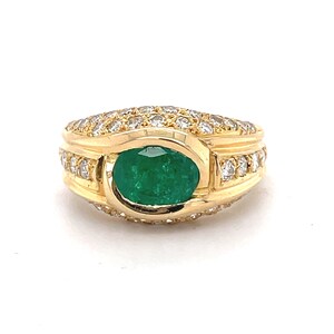 K&ouml;nnte beinhalten: Ein goldener Ring mit einem gro&szlig;en ovalen Smaragd, der von kleineren Diamanten umgeben ist. Der Ring ist in einer goldenen Band mit einem strukturierten Design gefasst.