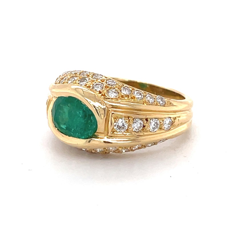 K&ouml;nnte beinhalten: Ein goldener Ring mit einem gro&szlig;en Smaragd, der von kleineren Diamanten umgeben ist. Der Ring hat eine breite Band mit einem strukturierten Design.