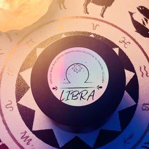 May include: A black circular candle tin with a white label featuring the text LIBRA, 100% SOY WAX, 8 OZ / 240 ML, HANDPOURED IN COCOA, FL, and STARLING STELLARY. The label displays a central graphic of a flower inside a semi-circle above a horizontal line.
