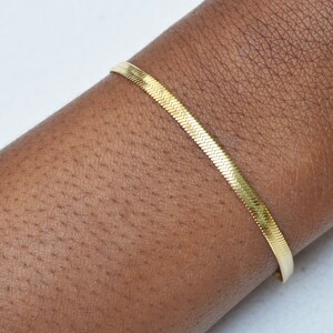 Peut inclure: Un bracelet chaîne à chevrons doré et fin, doté d'un design à maillons plats et flexibles qui repose sur la peau. La surface métallique reflète la lumière, mettant en valeur un motif en forme de V répété sur toute la longueur du bijou.