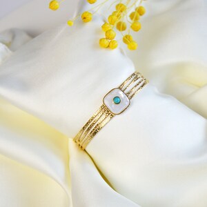 Może przedstawiać: Złota bransoletka z białym kwadratowym wisiorkiem ozdobionym turkusowym kamieniem. Bransoletka leży na białym tle z tkaniny.