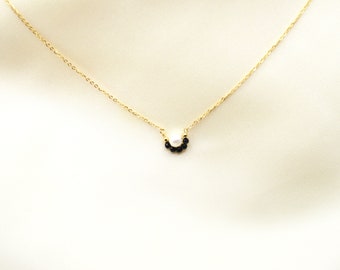 May include: A delicate gold chain necklace featuring a small, curved pendant. The pendant consists of a single white pearl centered above a row of five tiny, round black beads, creating a subtle, symmetrical arc design on the thin gold chain.