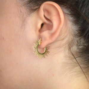 May include: Gold hoop earrings with a sunburst design. The earrings are worn in a person's ear.