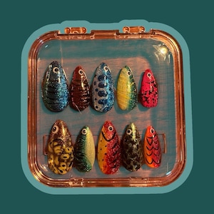 May include: A collection of ten fish-shaped nail art designs in a clear case. Each nail showcases a different pattern and colour scheme, including glitter and stripes. The designs are in a clear, rectangular case with a gold-coloured frame.