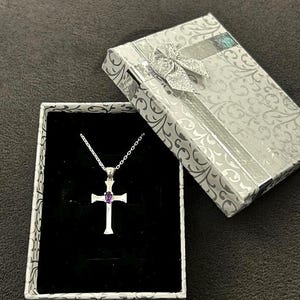Puede incluir: Un collar con una cruz de plata y una piedra preciosa púrpura en el centro, presentado en una caja de regalo blanca con un diseño plateado.