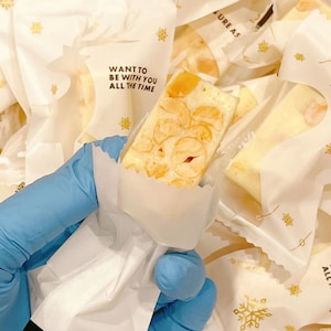 May include: A white paper bag with a gold snowflake design and the text "WANT TO BE WITH YOU ALL THE TIME" holds a piece of nougat candy with a light brown color and a white background.