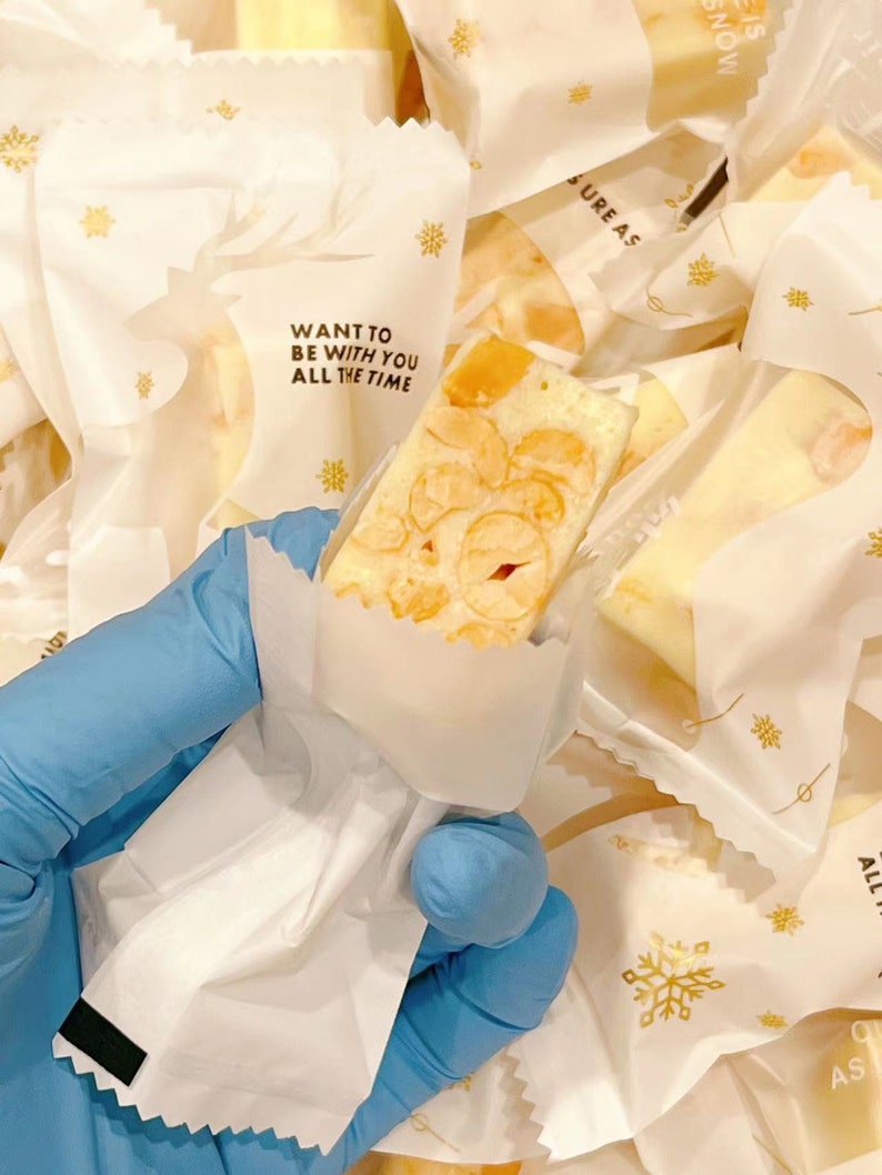 May include: A white paper bag with a gold snowflake design and the text "WANT TO BE WITH YOU ALL THE TIME" holds a piece of nougat candy with a light brown color and a white background.