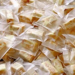 May include: A pile of individually wrapped nougat candy in clear plastic bags. The bags have various printed messages, including "Best Wishes", "Good Luck & Happiness", "Delicious", and "Thank You".