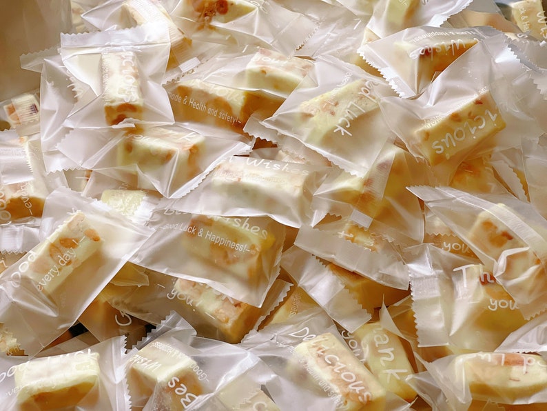 May include: A pile of individually wrapped nougat candy in clear plastic bags. The bags have various printed messages, including "Best Wishes", "Good Luck & Happiness", "Delicious", and "Thank You".