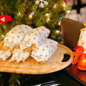 May include: A wooden serving tray with a handle holds five white gift bags with gold polka dots and gold ribbon bows. The bags are stacked on top of each other. A gingerbread man ornament and a lit candle are on a black surface in the background.