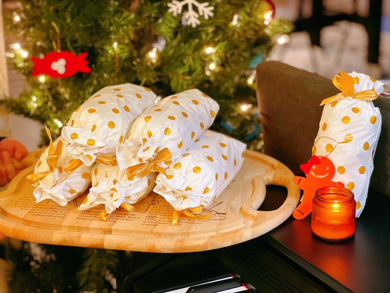 May include: A wooden serving tray with a handle holds five white gift bags with gold polka dots and gold ribbon bows. The bags are stacked on top of each other. A gingerbread man ornament and a lit candle are on a black surface in the background.
