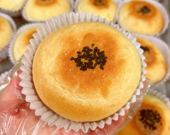 May include: A close-up of a single, round, golden brown bun with a small amount of black sesame seeds on top. The bun is held in a white paper liner and a gloved hand.