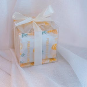 K&ouml;nnte beinhalten: Eine quadratische, durchsichtige Kunststoff-Geschenkbox mit einzeln verpackten Ananaskuchen. Die Verpackung zeigt ein gelbes Ananasmuster und den Text PINEAPPLE CAKE. Die Box ist mit einer cremefarbenen Satinschleife oben verziert.