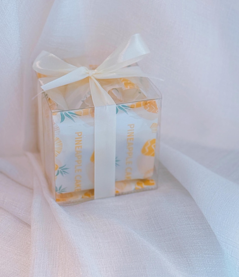 K&ouml;nnte beinhalten: Eine quadratische, durchsichtige Kunststoff-Geschenkbox mit einzeln verpackten Ananaskuchen. Die Verpackung zeigt ein gelbes Ananasmuster und den Text PINEAPPLE CAKE. Die Box ist mit einer cremefarbenen Satinschleife oben verziert.