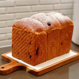 May include: A loaf of freshly baked bread with a dusting of powdered sugar. The bread is brown and has a slightly sweet, raisin-like appearance. It is sitting on a wooden cutting board with a white surface.